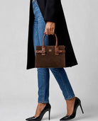 Brown structured handbag carried by hand with black coat, blue jeans and black heels.