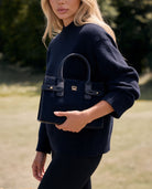 Navy structured handbag carried with a navy knit sweater and black trousers in an outdoor setting.