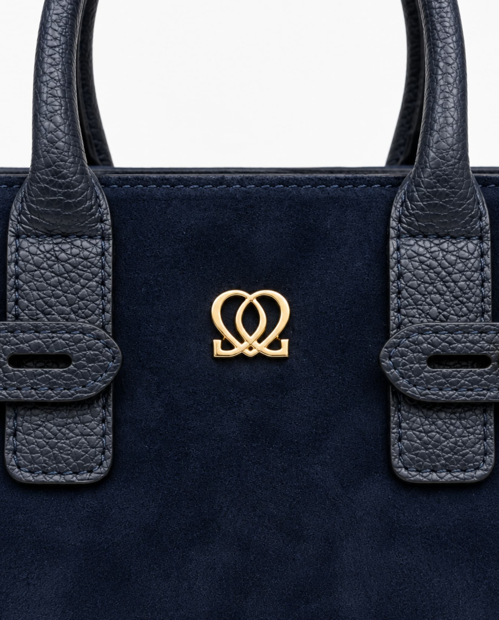 Close-up of navy handbag showing suede-like material, textured handle details and centered gold-tone emblem.