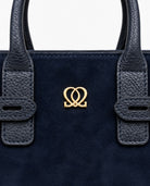 Close-up of navy handbag showing suede-like material, textured handle details and centered gold-tone emblem.